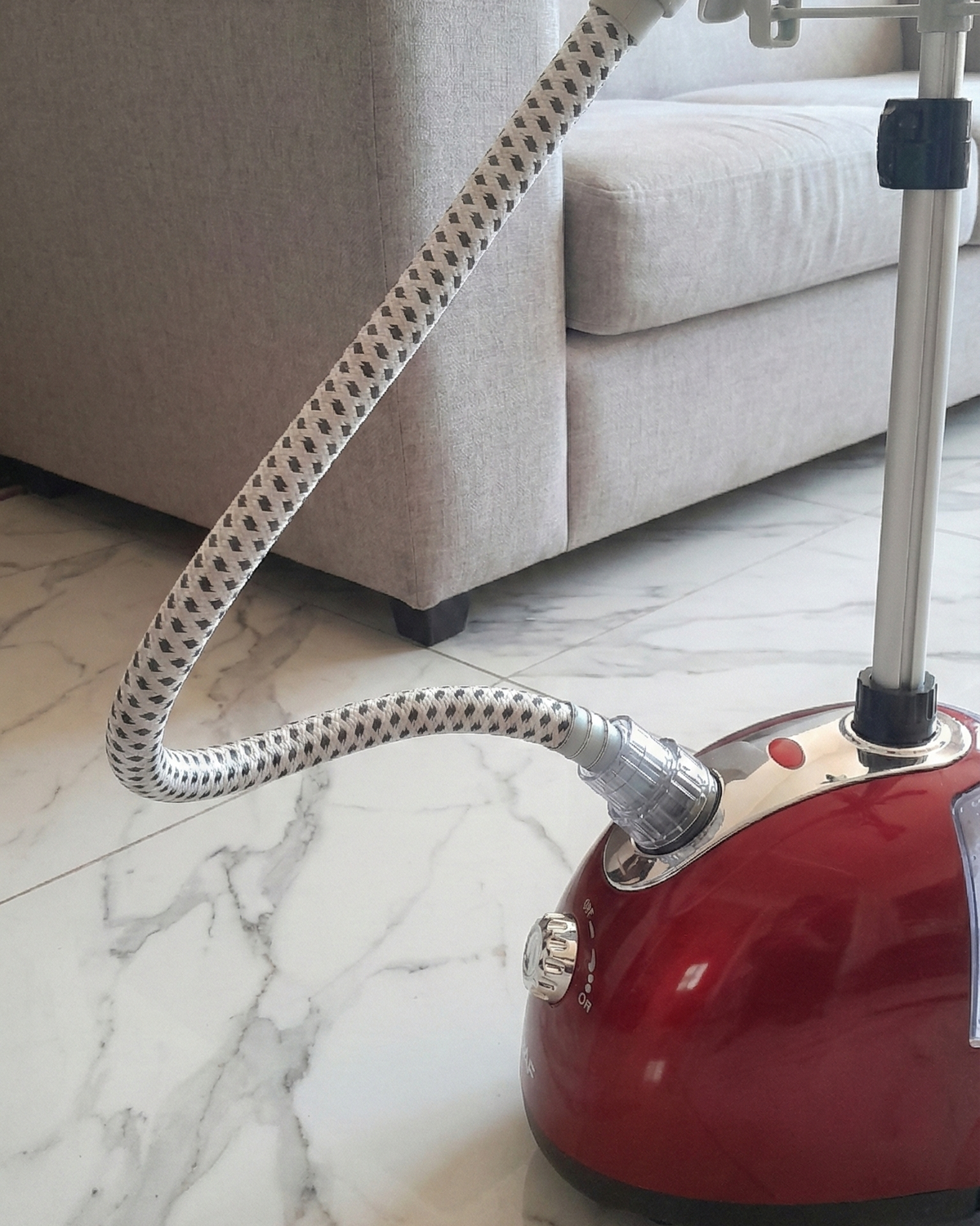 Red garment steamer with flexible hose on a marble floor, with a gray sofa in the background.